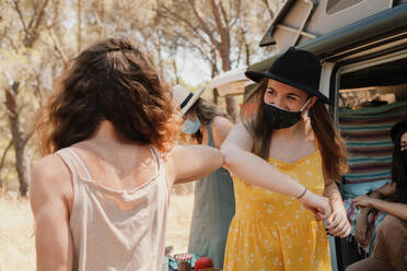 Friendly women in protective masks standing near van in forest and bumping elbows while greeting each other during coronavirus epidemic - ADSF18357