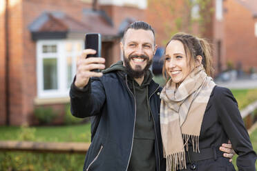 Lächelnder Mann, der ein Selfie mit seiner Freundin macht, während er vor seinem neuen Haus steht - WPEF03708