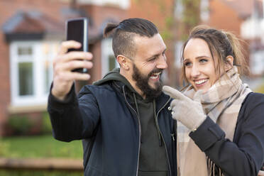 Junge Frau gestikuliert, während sie ein Selfie mit ihrem fröhlichen Freund vor einem neuen Haus macht - WPEF03709