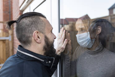 Mann sieht seine Freundin mit Gesichtsschutzmaske durch das Fensterglas während COVID-19 - WPEF03713