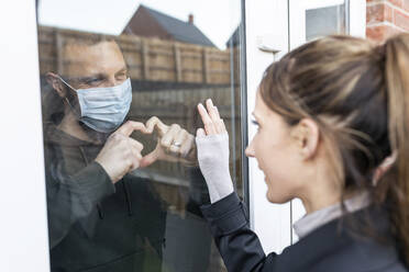 Freund zeigt seiner Freundin durch das Fensterglas eine herzförmige Geste, während er zu Hause unter Quarantäne steht - WPEF03716