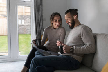 Smiling young couple having tea while using laptop on sofa at home - WPEF03717