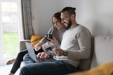 Lächelndes Paar beim Online-Shopping mit dem Laptop, während sie zu Hause Tee trinken - WPEF03719