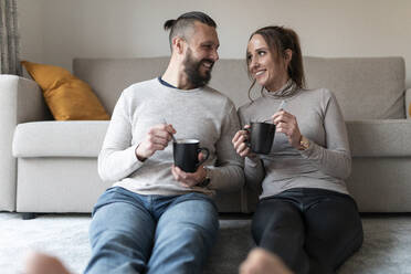 Smiling couple looking at each other while sitting on carpet in living room at home - WPEF03727