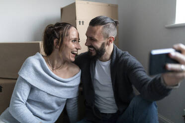 Cheerful couple looking face to face while taking selfie at new home - WPEF03732