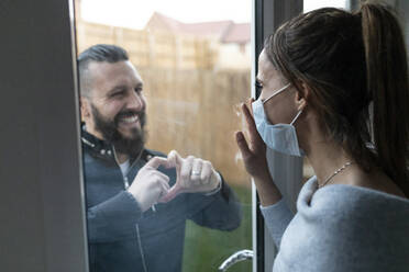 Freund zeigt seiner Freundin durch das Fensterglas eine herzförmige Geste während der COVID-19-Krise - WPEF03735