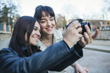 Smiling woman standing by female friend taking photograph with DSLR camera - MGRF00066