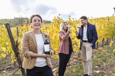 Female entrepreneur holding wine bottle while clients standing and drinking during autumn - EIF00350