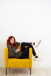 Redhead woman using mobile phone while resting on arm chair at office - GIOF09848
