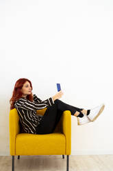 Young woman taking selfie through mobile phone while sitting on armchair at office - GIOF09849