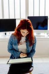 Junge Geschäftsfrau, die ein digitales Tablet benutzt, während sie im Büro auf einem Hocker sitzt - GIOF09850