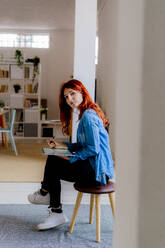 Rothaarige Geschäftsfrau, die ein digitales Tablet benutzt, während sie auf einem Hocker im Büro sitzt - GIOF09852
