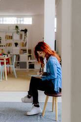 Junge Geschäftsfrau, die im Büro auf einem Hocker sitzend an einem digitalen Tablet arbeitet - GIOF09853