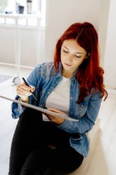 Rothaarige Geschäftsfrau arbeitet am digitalen Tablet, während sie im Büro auf dem Boden sitzt - GIOF09858