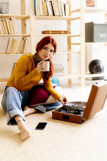 Junge Frau trinkt Kaffee, während sie zu Hause am Plattenspieler sitzt - GIOF09872