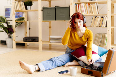 Redhead young wearing headphones using turntable while sitting at home - GIOF09874
