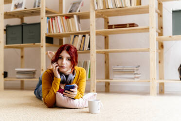 Woman with coffee cup using mobile phone while lying on carpet at home - GIOF09878