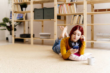 Young woman using mobile phone while lying on carpet at home - GIOF09880