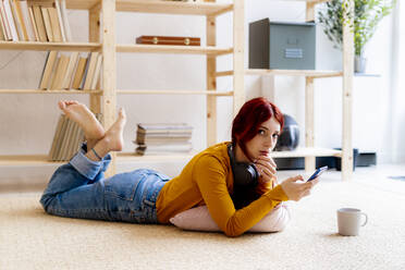 Rothaarige Frau, die ein Mobiltelefon benutzt, während sie zu Hause auf dem Teppich liegt - GIOF09881