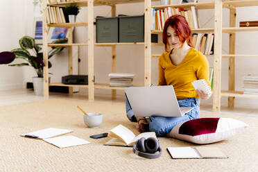 Young woman using laptop while sitting against bookshelf at home - GIOF09882