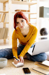 Young woman with laptop on lap writing in note pad while studying at home - GIOF09883