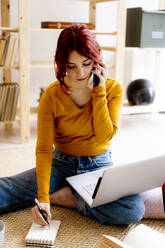 Junge Frau mit Laptop, die zu Hause sitzt und mit ihrem Mobiltelefon spricht - GIOF09886