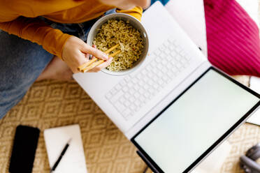 Young woman eating noodles while studying through laptop sitting at home - GIOF09889