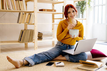 Lächelnde Frau beim Essen und Lernen am Laptop zu Hause sitzend - GIOF09891