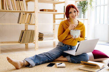 Smiling woman having food while studying through laptop sitting at home - GIOF09891