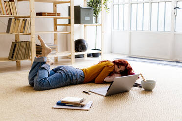 Rothaarige Frau mit Buch und Laptop auf Teppich liegend zu Hause - GIOF09893