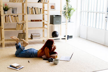 Thoughtful woman with laptop looking away while lying on carpet at home - GIOF09899