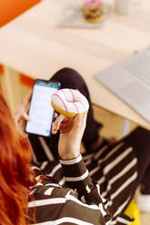 Businesswoman holding doughnut while using mobile phone sitting at office - GIOF09907