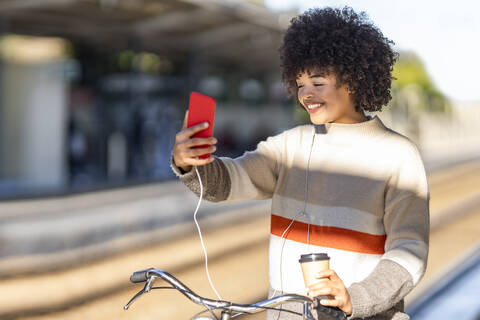 Young woman taking selfie while standing with bicycle and reusable cup at railroad station stock photo