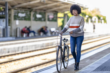 Young woman walking with bicycle while day dreaming at railroad station platform - GGGF00232