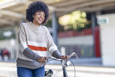 Cheerful young woman wheeling bicycle while looking away at railroad station - GGGF00233