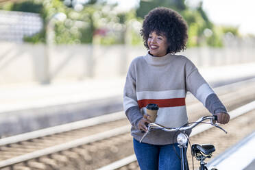 Nachdenkliche junge Frau mit wiederverwendbarem Becher auf dem Fahrrad am Bahnhof - GGGF00235