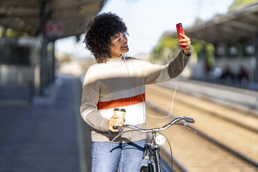 Smiling young woman with bicycle taking selfie while listening music at station - GGGF00237