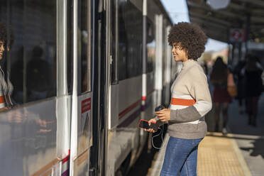 Glücklicher junger weiblicher Fahrgast beim Einsteigen in den Zug am Bahnhof an einem sonnigen Tag - GGGF00241