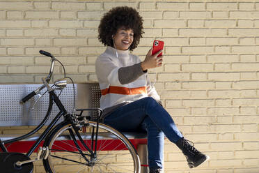 Glückliche Frau, die ein Selfie macht, während sie auf einer Bank mit einem Fahrrad vor einer Mauer sitzt - GGGF00242