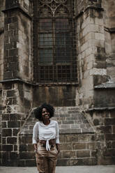 Smiling woman with hands in pockets standing against Barcelona Cathedral, Barcelona, Spain - GMLF00852