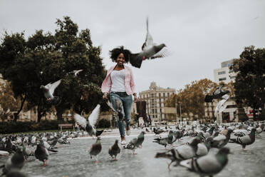 Cheerful woman running amidst flock on pigeons at park in city - GMLF00855