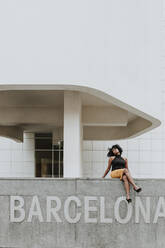 Carefree woman sitting on retaining wall with Barcelona text - GMLF00869