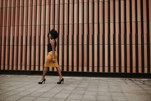 Mature woman walking on footpath by striped wall - GMLF00870