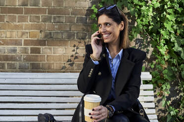 Smiling businesswoman with bamboo cup talking on smart phone while sitting on bench - VEGF03215