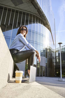 Lächelnde Geschäftsfrau sitzt auf einer Bank neben einem Bürogebäude - VEGF03222