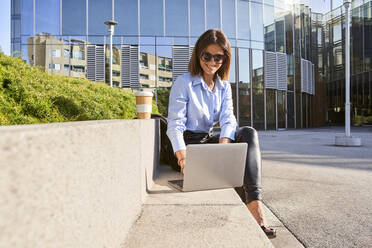 Happy female entrepreneur using laptop while sitting on bench against office building during sunny day - VEGF03223