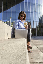 Smiling businesswoman using laptop while holding bamboo cup against office building - VEGF03224
