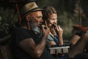 Smiling friends eating vegetable while sitting on bench outdoors - MJRF00307