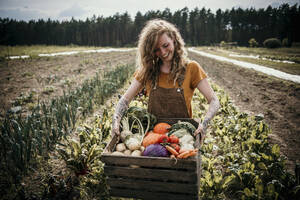 Lächelnder Landarbeiter, der eine Gemüsekiste trägt, während er auf einem Bauernhof steht - MJRF00311