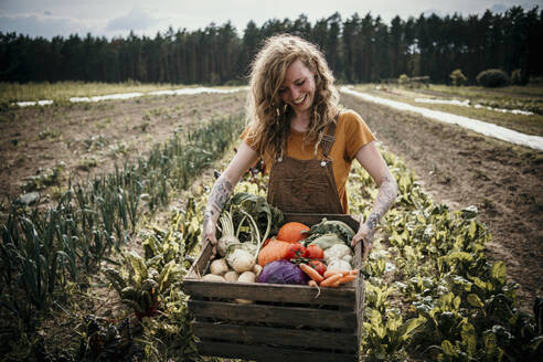 Smiling farm worker carrying vegetable box while standing at farm - MJRF00311